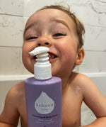 A close up of a child in a bath and holding the Kokoso softly scented organic baby hair body & wash bottle. Bottle available at Babipur.