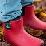 A close up of a child wearing the Little Green Radicals ankle wellington boots in red on wet leaves showing the black ankle elasticated cuff detail