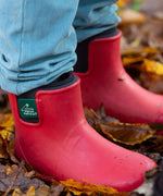 A close up of a child wearing the Little Green Radicals ankle wellington boots in red on wet leaves showing the black ankle elasticated cuff detail