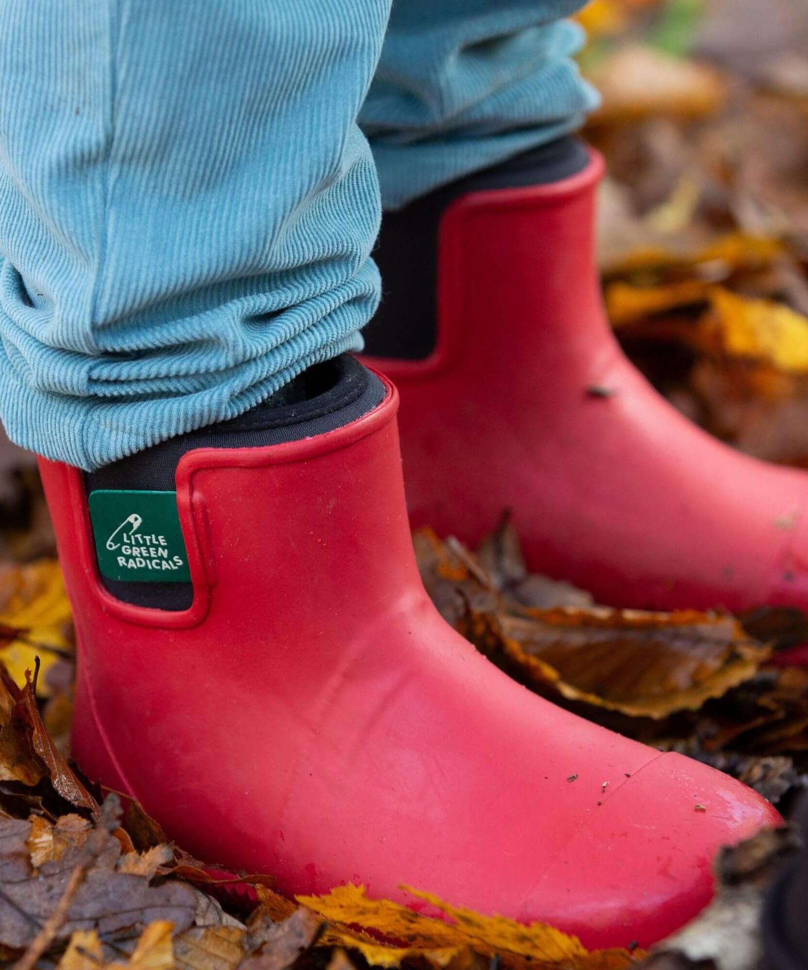 A close up of a child wearing the Little Green Radicals ankle wellington boots in red on wet leaves showing the black ankle elasticated cuff detail