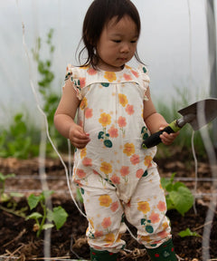 Small child wearing Little Green Radicals kids flora frill summer romper dress in cream with a repeating floral design in a garden