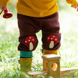 A close up of a child standing in grass and wearing the Little Green Radicals knee patch joggers showing the toadstool knee patch applique detail