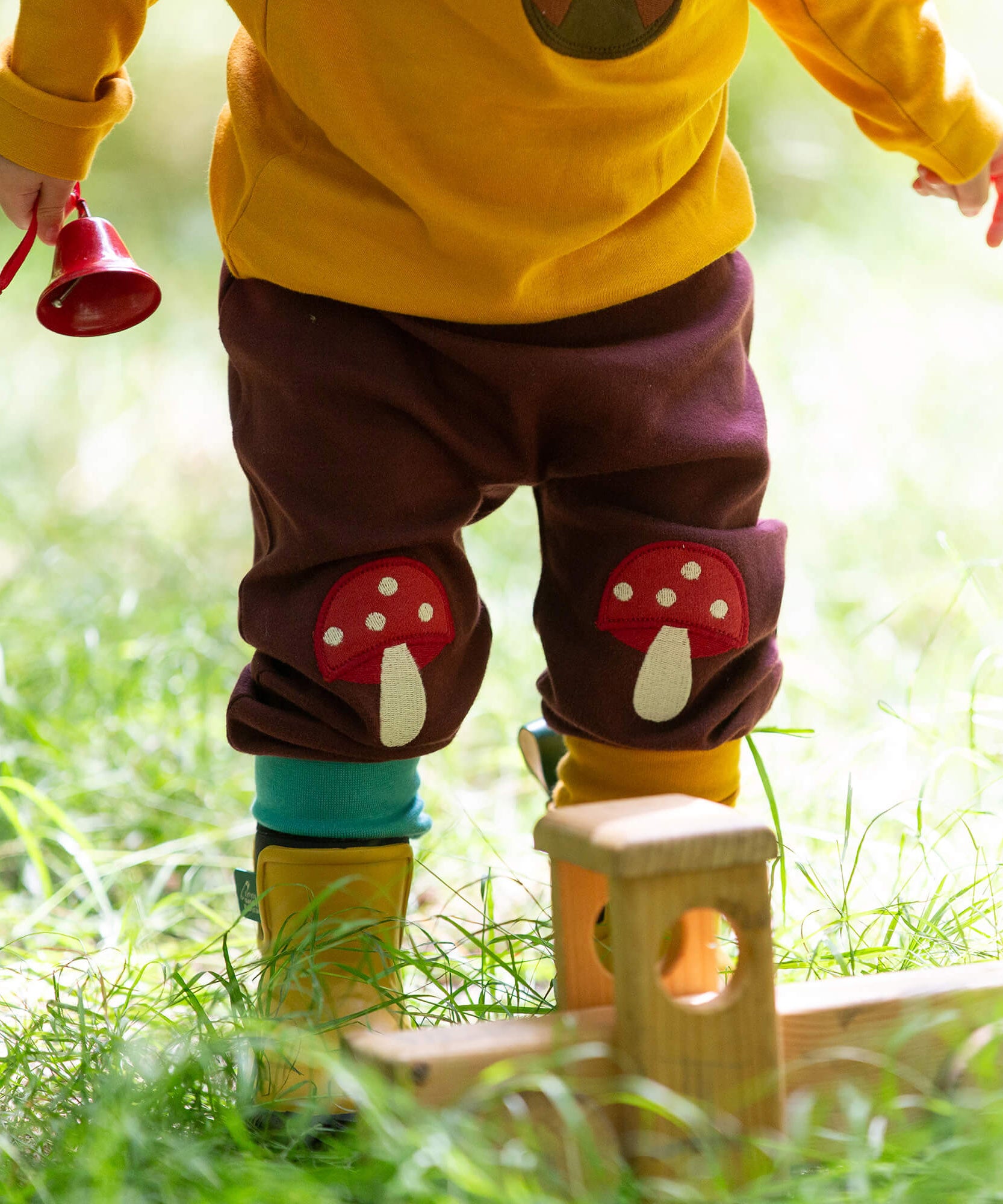 A close up of a child standing in grass and wearing the Little Green Radicals knee patch joggers showing the toadstool knee patch applique detail
