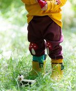 A close up of a child standing in grass and wearing the Little Green Radicals knee patch joggers showing the toadstool knee patch applique detail and the light blue and yellow cuff trim.