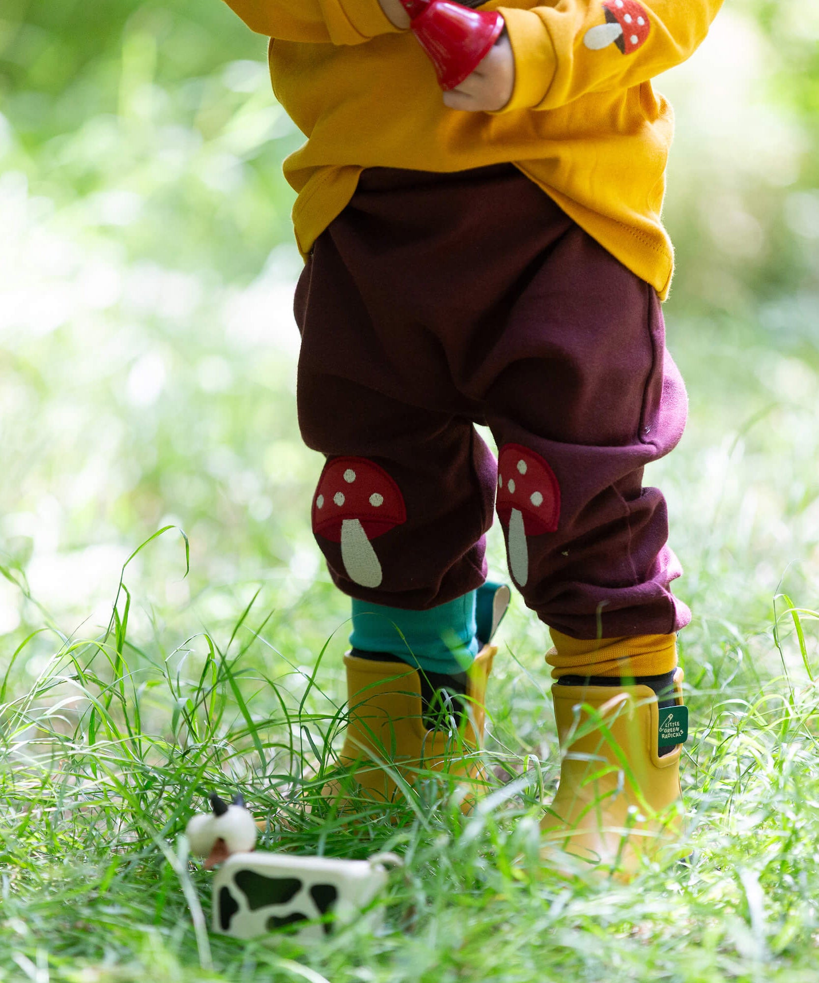 A close up of a child standing in grass and wearing the Little Green Radicals knee patch joggers showing the toadstool knee patch applique detail and the light blue and yellow cuff trim.