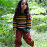 A child standing on grass with trees in the background and wearing the Little Green Radicals rainbow waffle snuggly knitted jumper.