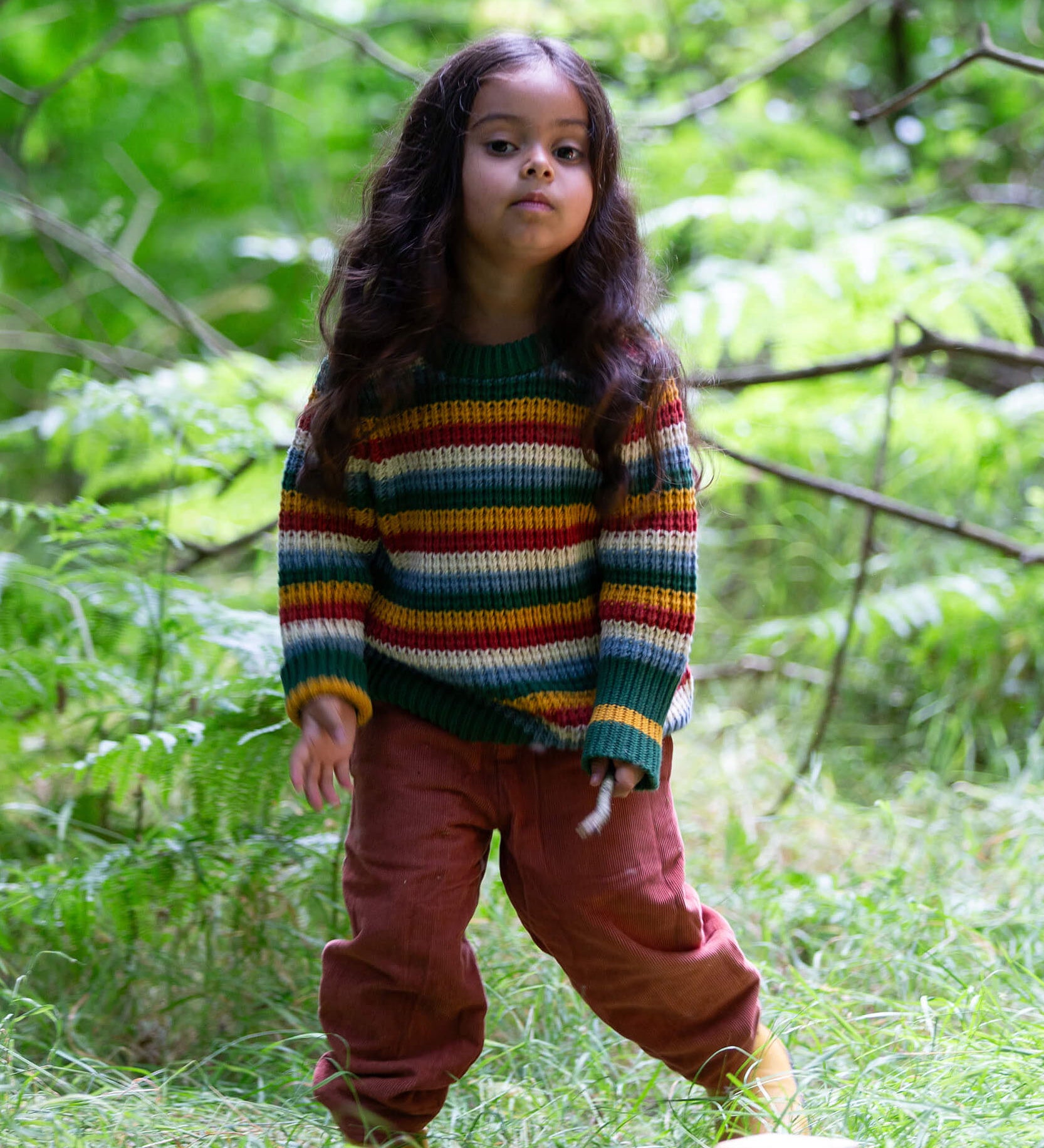 A child standing on grass with trees in the background and wearing the Little Green Radicals rainbow waffle snuggly knitted jumper.