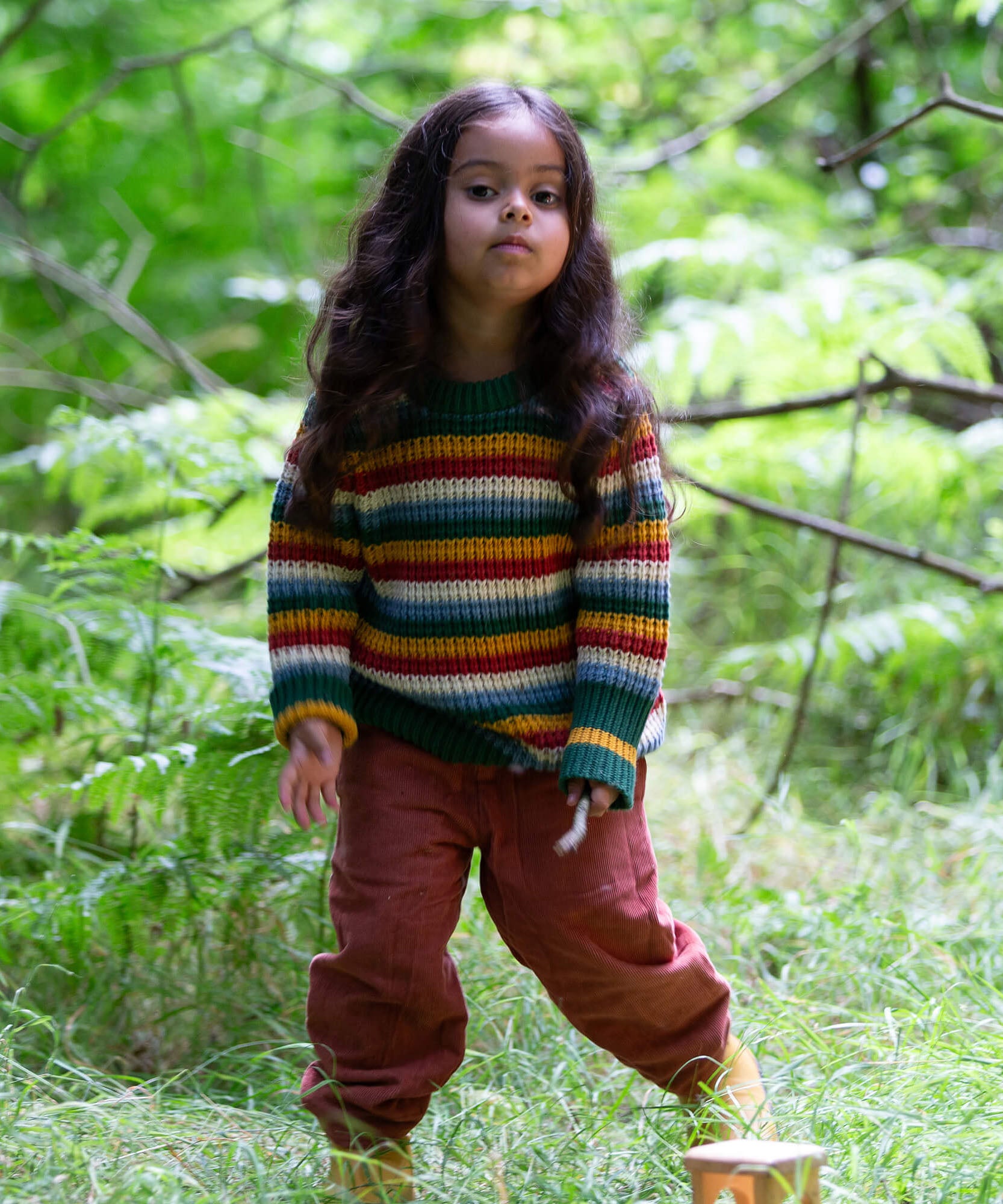 A child standing on grass with trees in the background and wearing the Little Green Radicals rainbow waffle snuggly knitted jumper.
