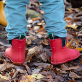 A child standing on wet leaves and showing the back detail of the Little Green Radicals ankle wellington boots in red