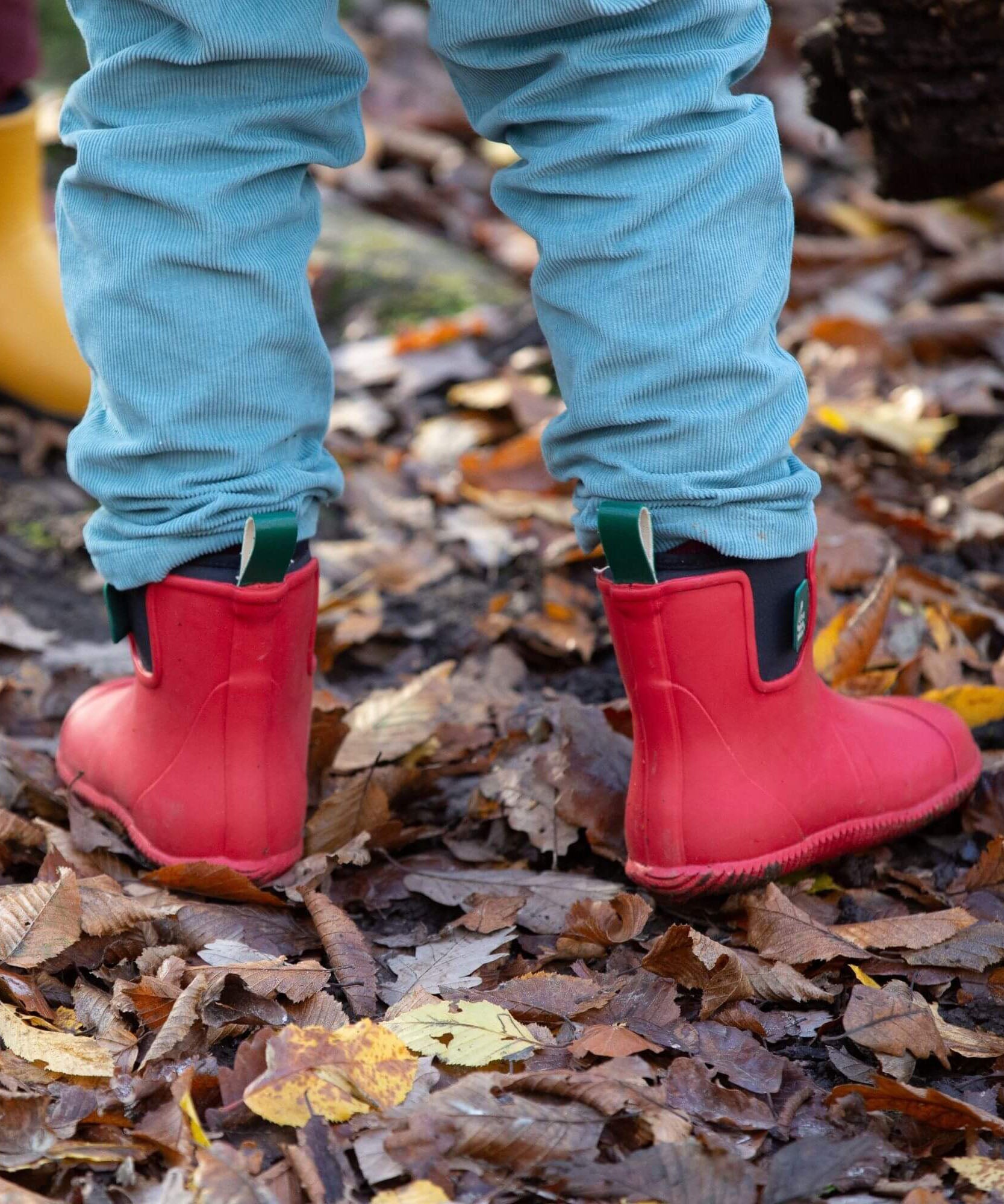 A child standing on wet leaves and showing the back detail of the Little Green Radicals ankle wellington boots in red