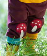 A close up of a child wearing the dark maroon Little Green Radicals knee patch joggers showing the toadstool knee patch applique detail