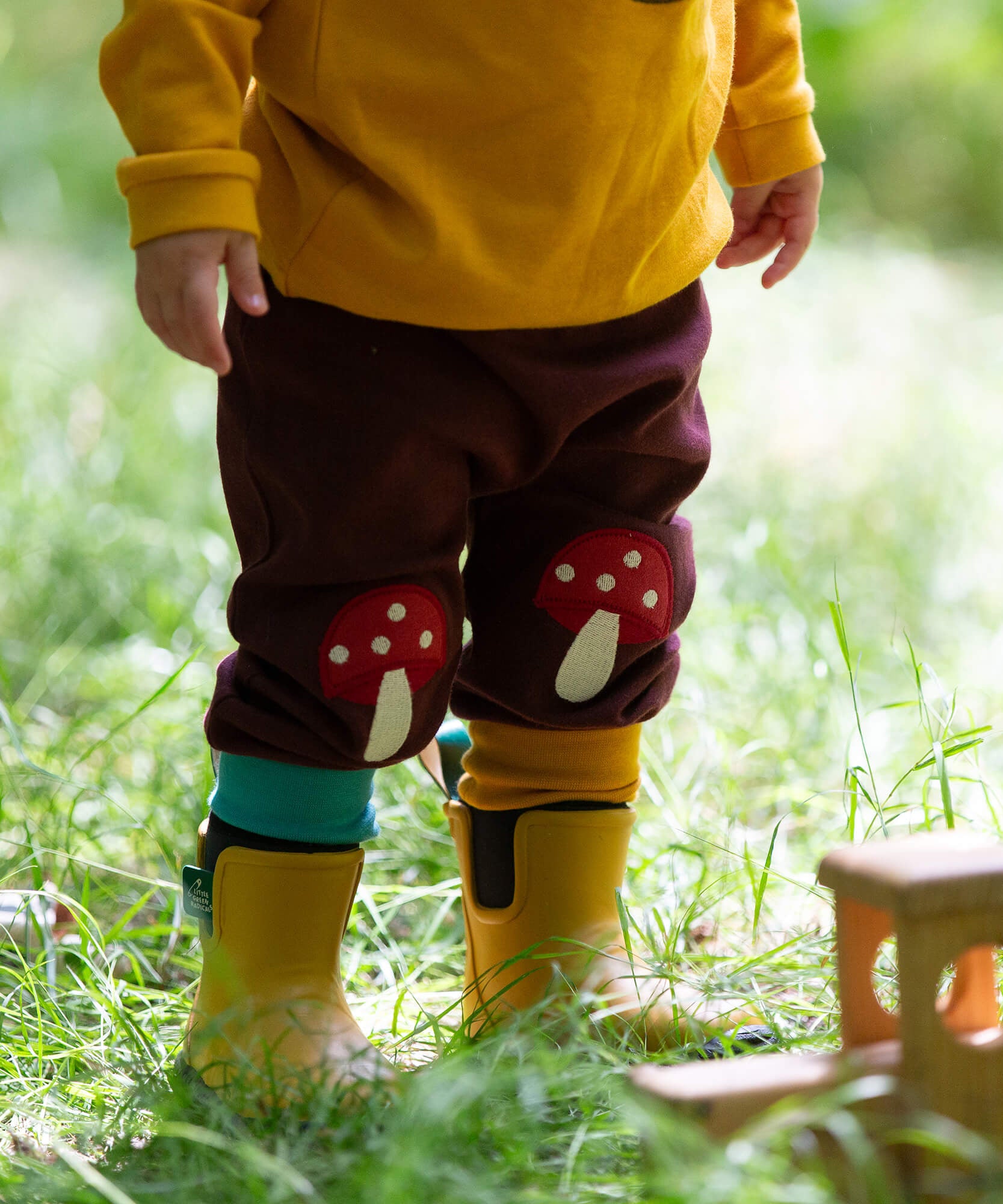 A close up of a child standing in grass and wearing the Little Green Radicals knee patch joggers showing the light blue and yellow leg cuff trim.