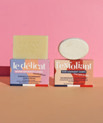 The Lamazuna Solid Exfoliant Bar and the Delicate Gentle Soap Bar, stood on top of their packaging boxes, on a pink and peach background