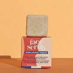 The Lamazuna Solid Face Scrub Bar stood on top of its packaging box, on a yellow and orange background