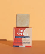 The Lamazuna Solid Face Scrub Bar stood on top of its packaging box, on a yellow and orange background