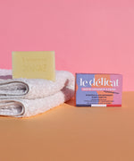 The Lamazuna Sensitive Skin Soap Bar is stood on top of some folded white fluffy towels, with the packaging box text to the towels, on a pink and orange background
