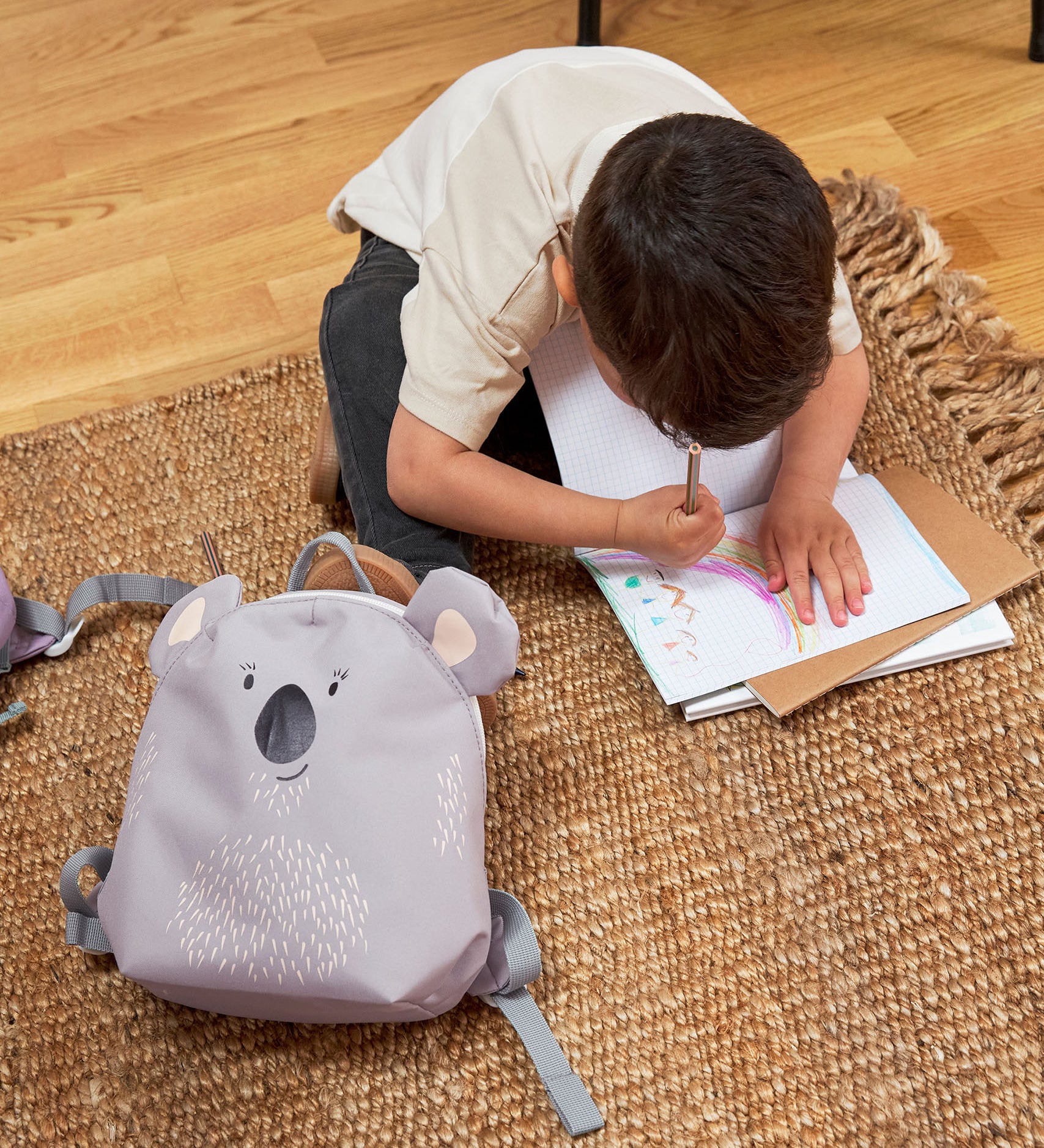 A child sitting on a rug, drawing next to the Lassig koala tiny backpack.