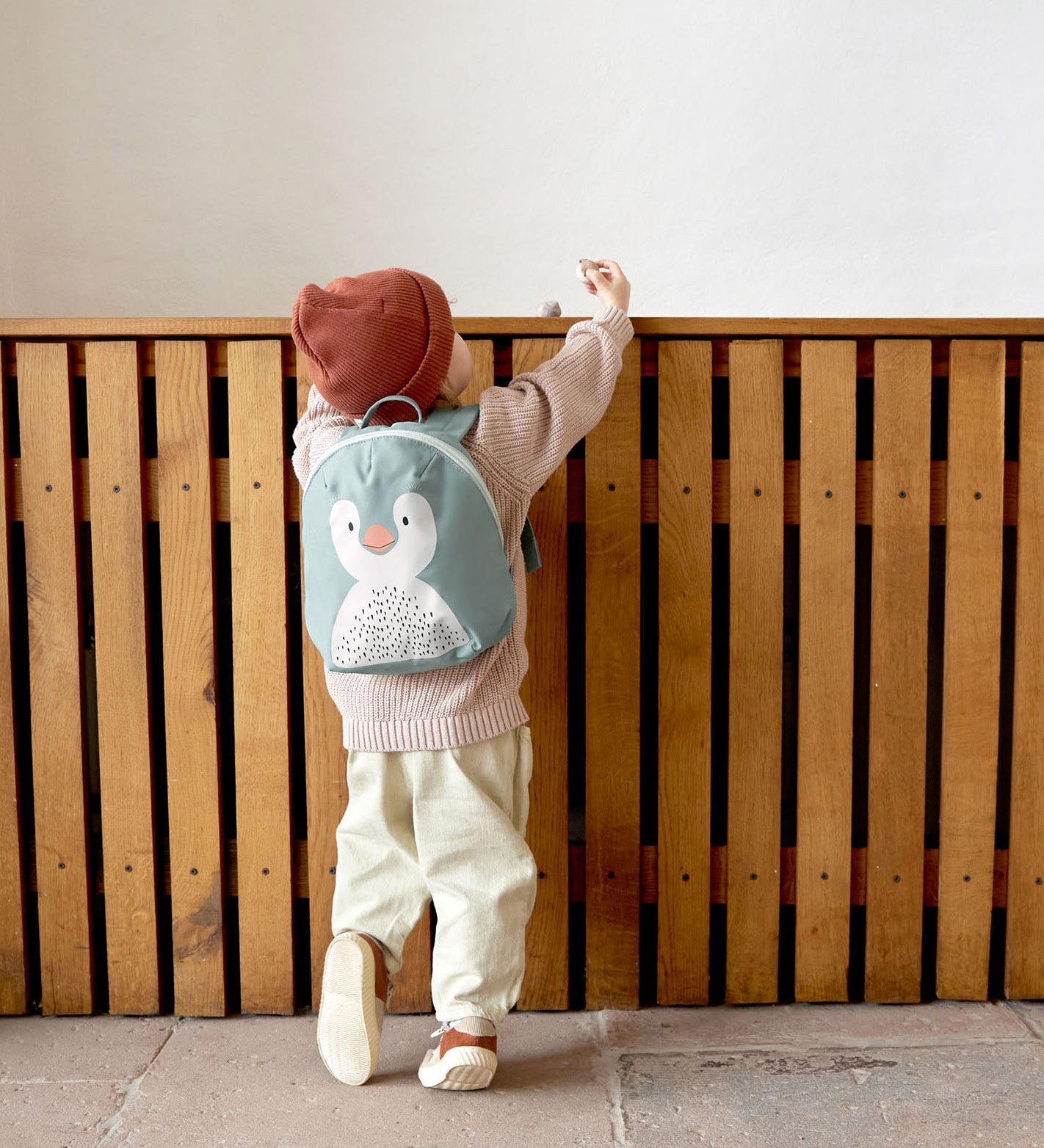 A small child standing near a wooden fence and wearing the Lassig kids tiny backpack in penguin 
