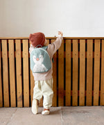 A small child standing near a wooden fence and wearing the Lassig kids tiny backpack in penguin 