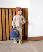 A small child standing near a wooden fence and holding the Lassig kids tiny backpack in whale by the top handle 