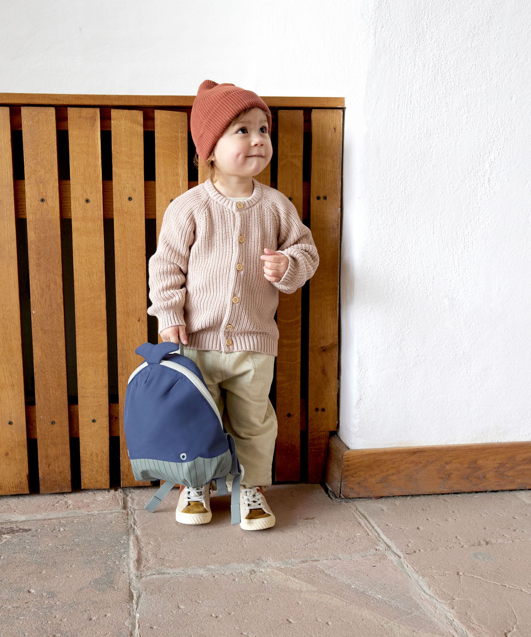 A small child standing near a wooden fence and holding the Lassig kids tiny backpack in whale by the top handle 