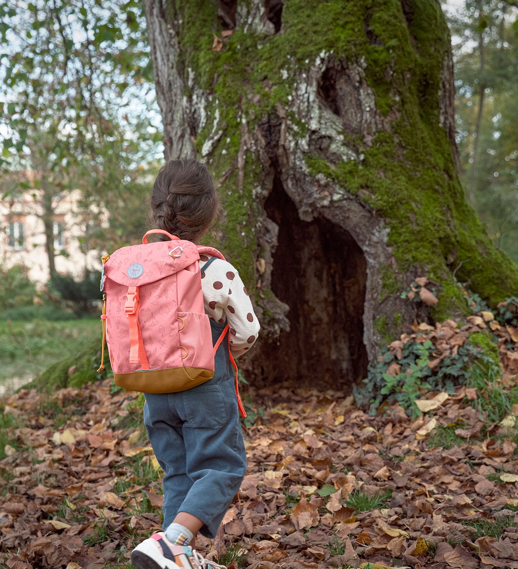 A child walking through leaves near a big tree and wearing the Lassig kids mini advetnure backpack in rose.