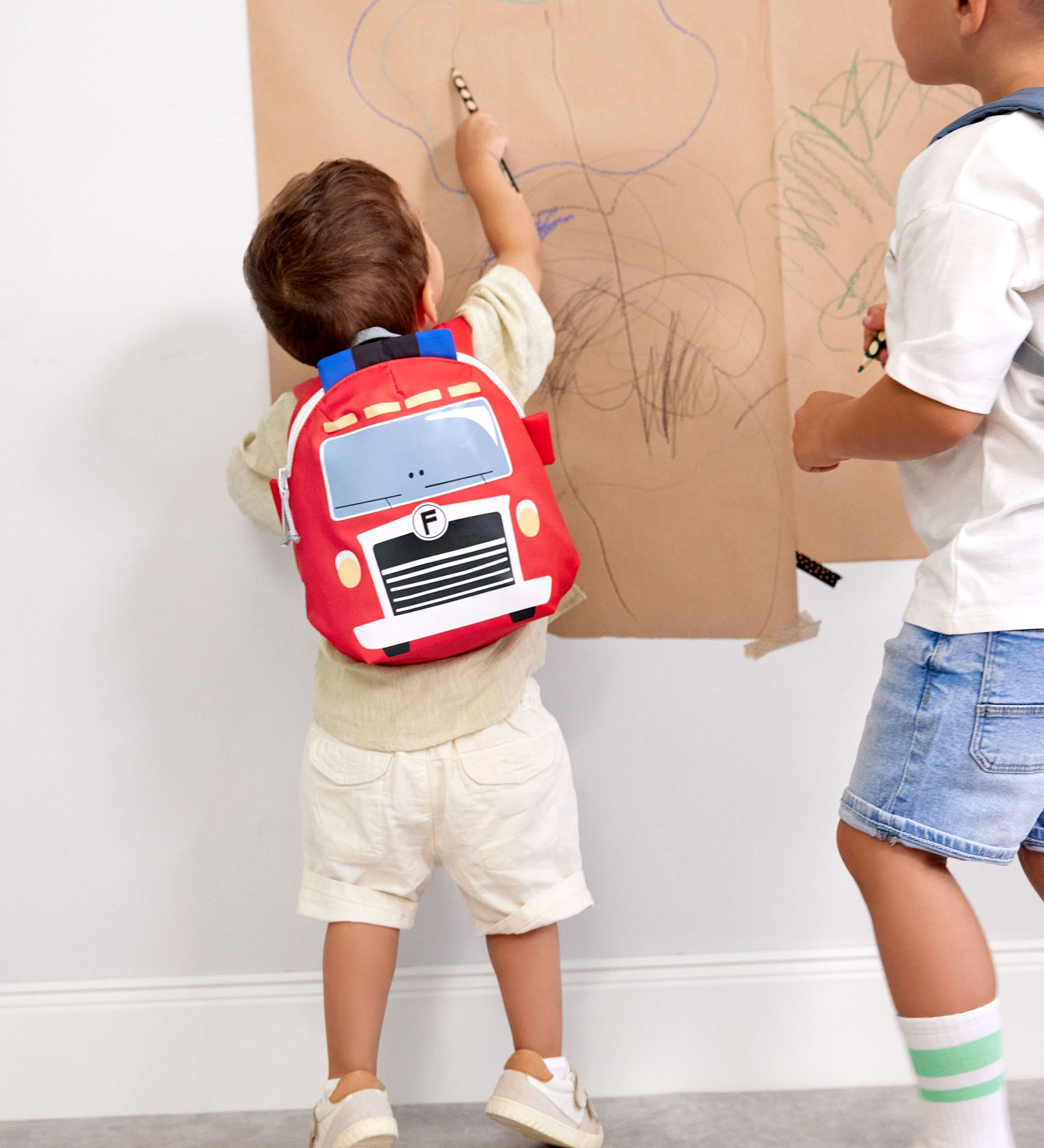 A child drawing on paper in a playroom and wearing the Lassig kids tiny fire engine backpack