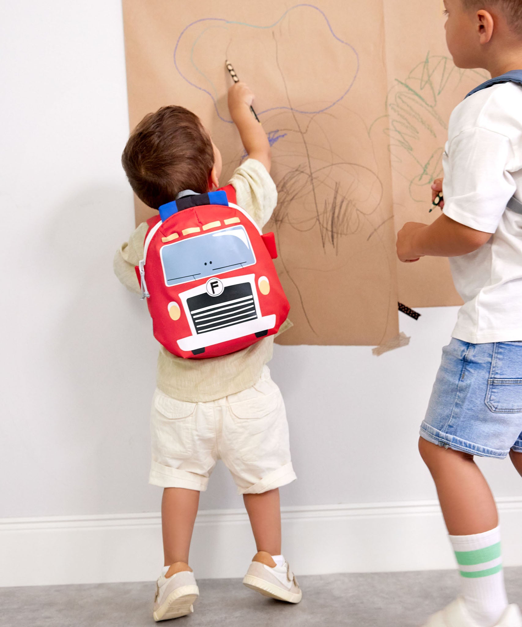 A child drawing on paper in a playroom and wearing the Lassig kids tiny fire engine backpack