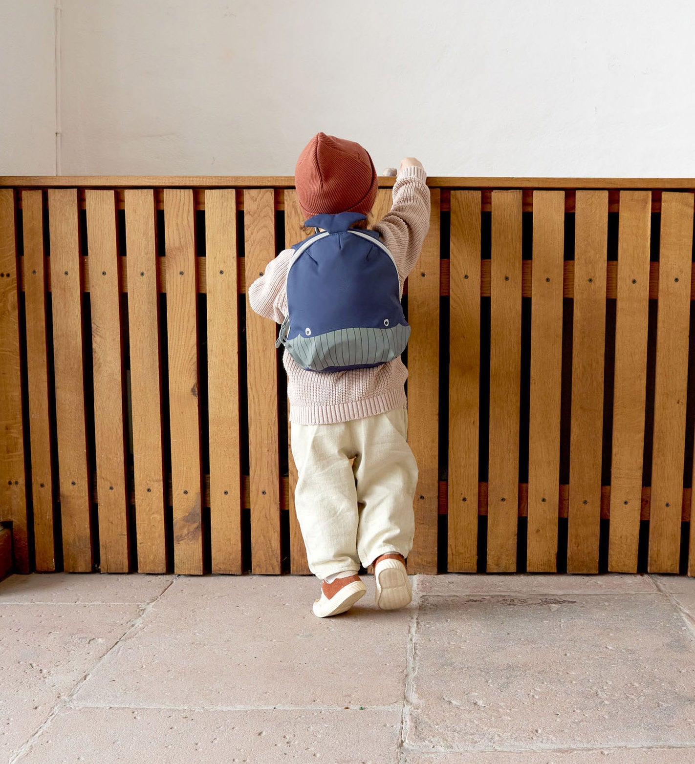 A small child standing near a wooden fence and wearing the Lassig kids tiny backpack in whale 