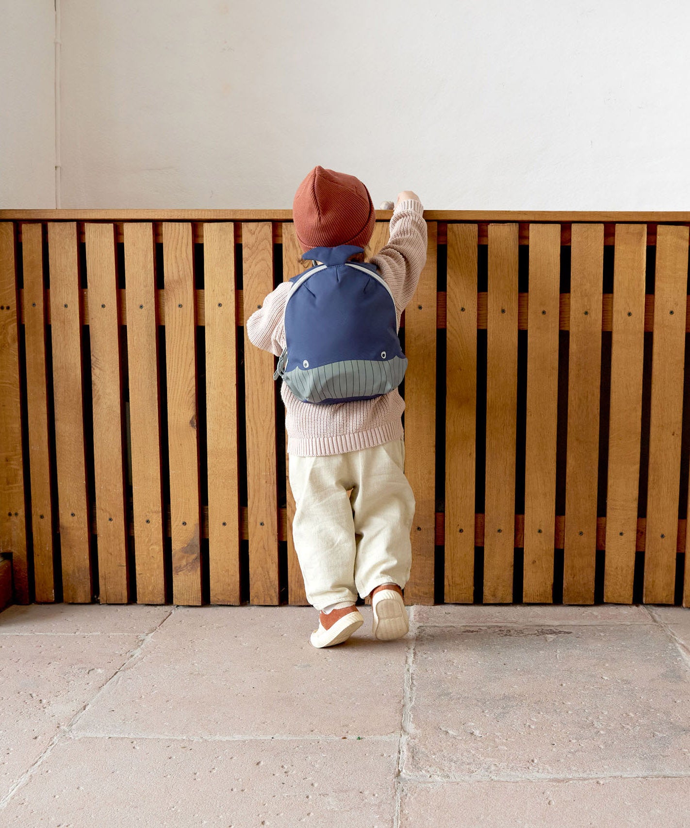 A small child standing near a wooden fence and wearing the Lassig kids tiny backpack in whale 