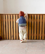 A small child standing near a wooden fence and wearing the Lassig kids tiny backpack in whale 