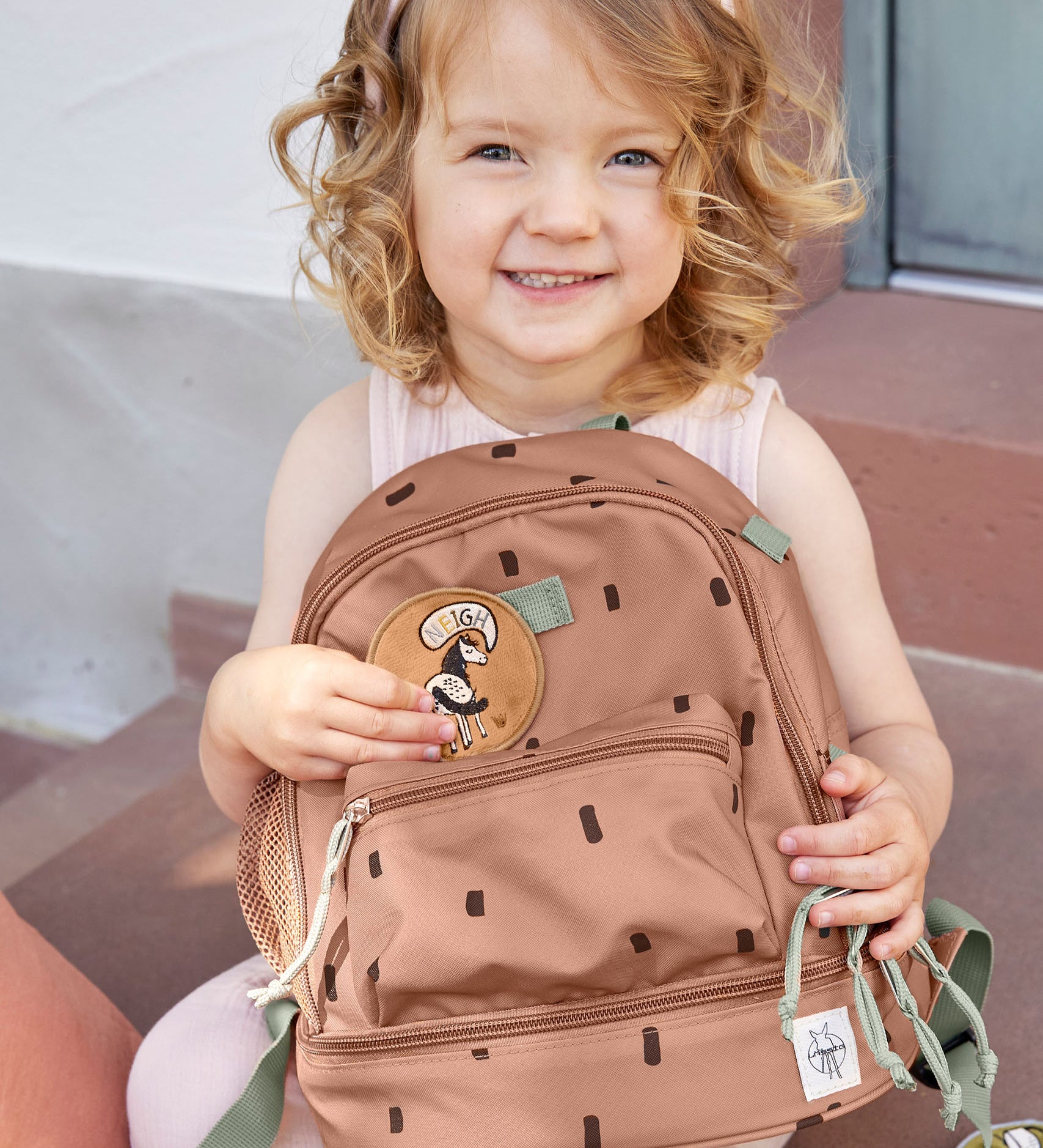 A child sitting outside and holding the Lassig kids mini happy prints Caramel pattern backpack. A pale brown backpack with a cute horse patch on the front panel. 