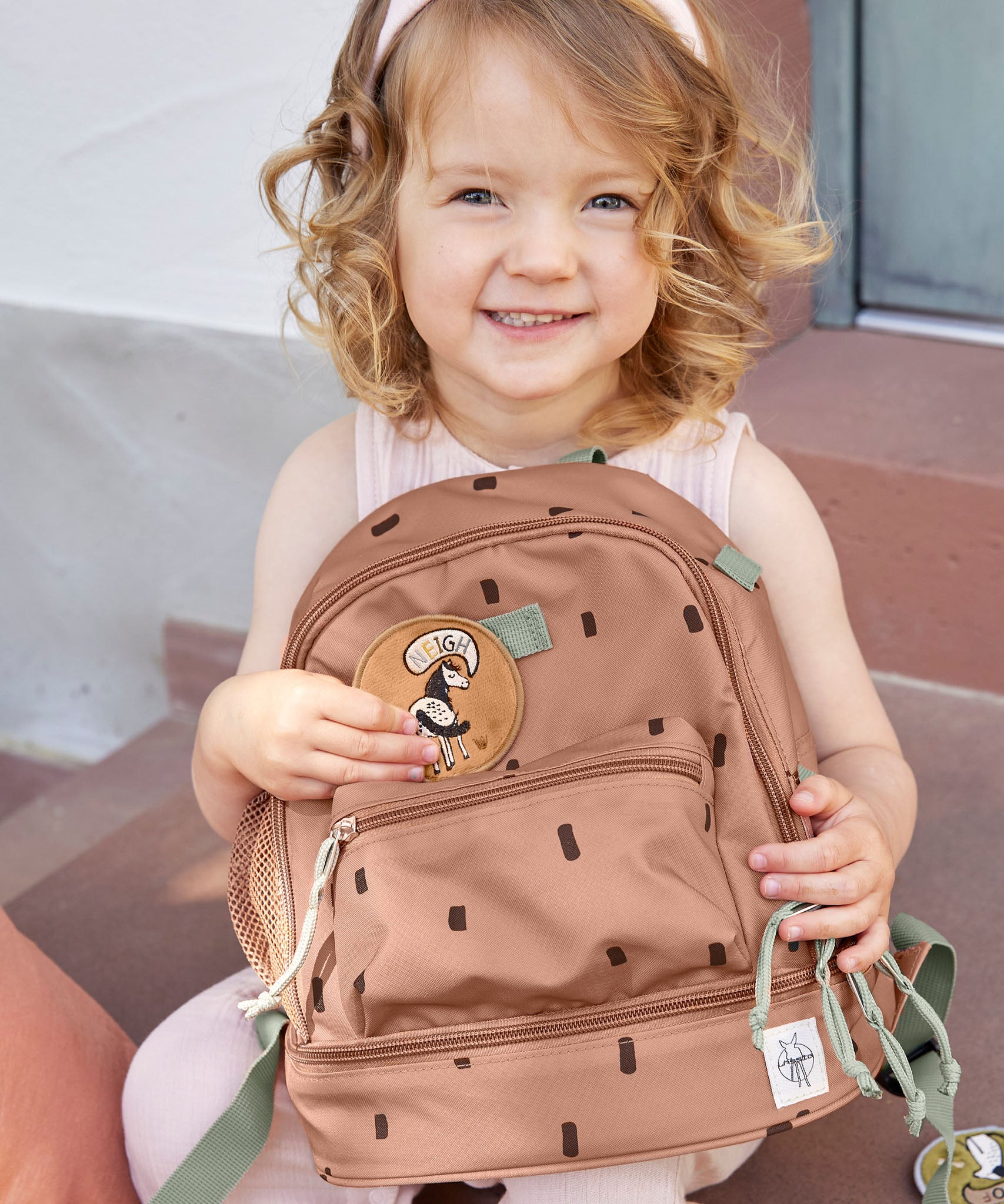 A child sitting outside and holding the Lassig kids mini happy prints Caramel pattern backpack. A pale brown backpack with a cute horse patch on the front panel. 