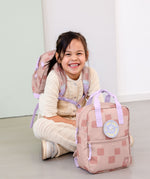 A child sitting on the floor and wearing the Lassig kids mini pattern party rose pattern backpack with another in front. 