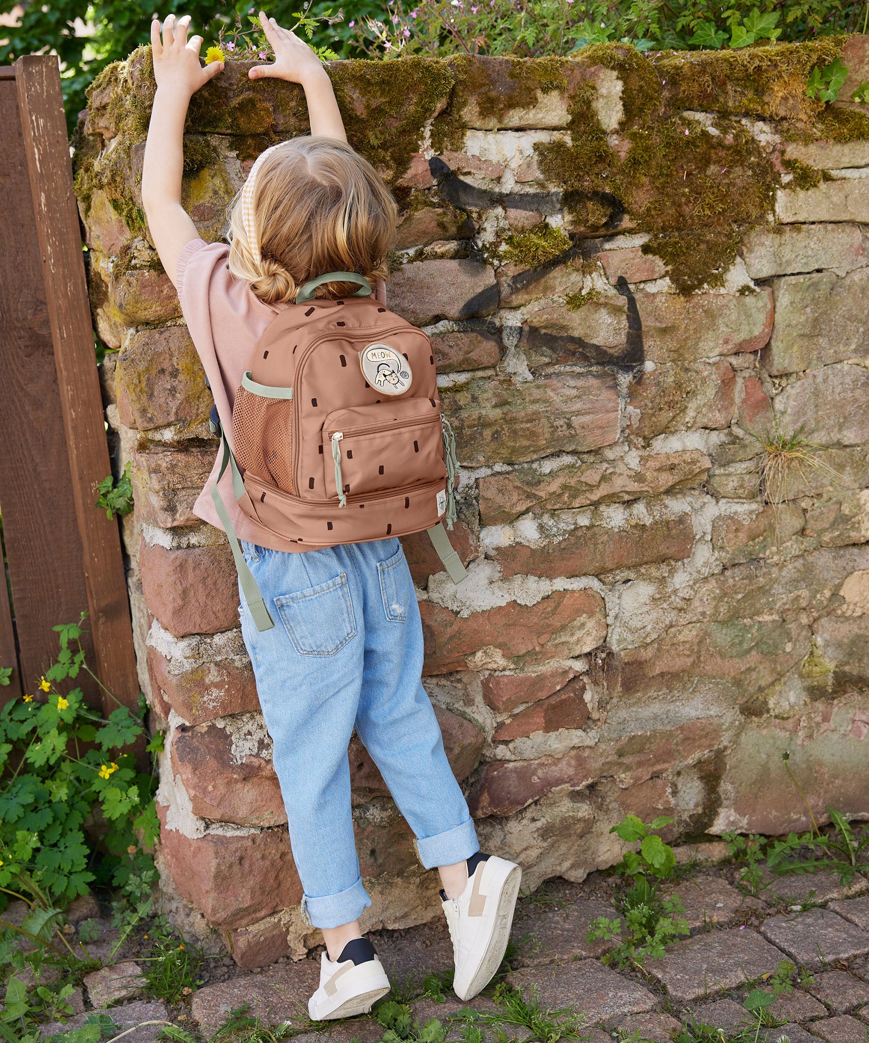 A child standing by a wall and wearing the Lassig kids mini happy prints caramel backpack