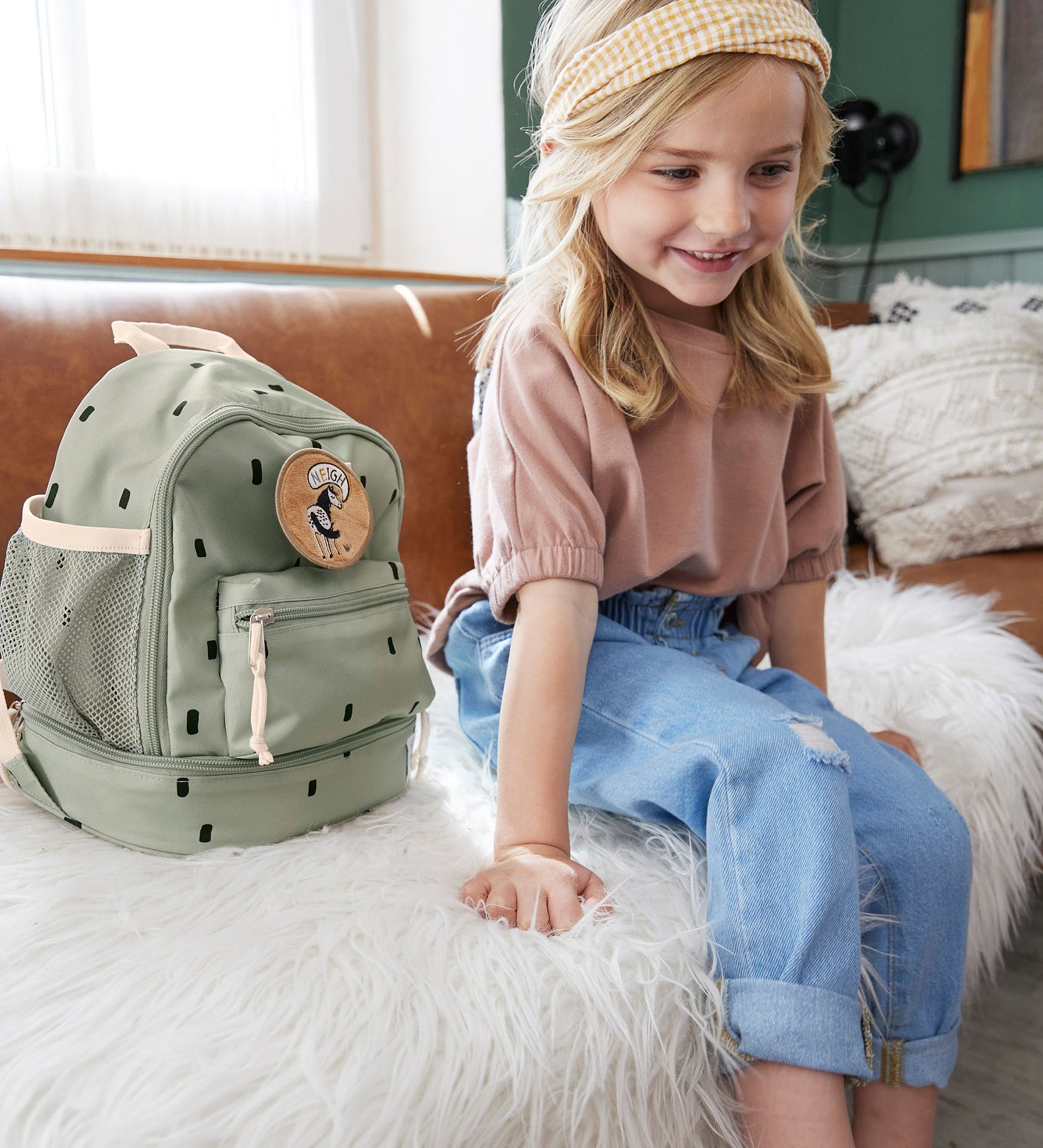 A child sitting on a couch next to the Lassig kids mini happy prints olive backpack