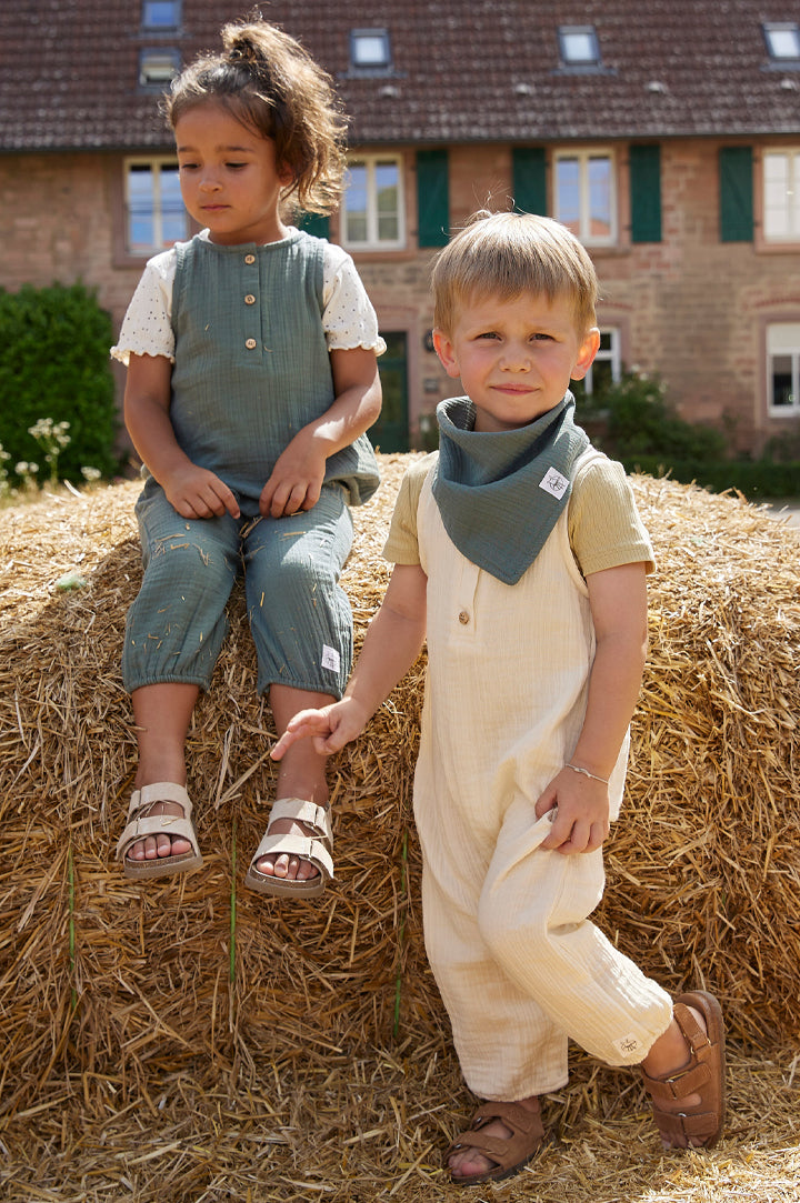 Two children sat outdoors on a haystack wearing GOTS organic cotton muslin dungarees by Lassig