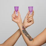 Two people holding a small and large purple Silicone, BPA free and latex free Ruby Cup in between their fingers. The image shows their arms crossing over and is on a light grey background