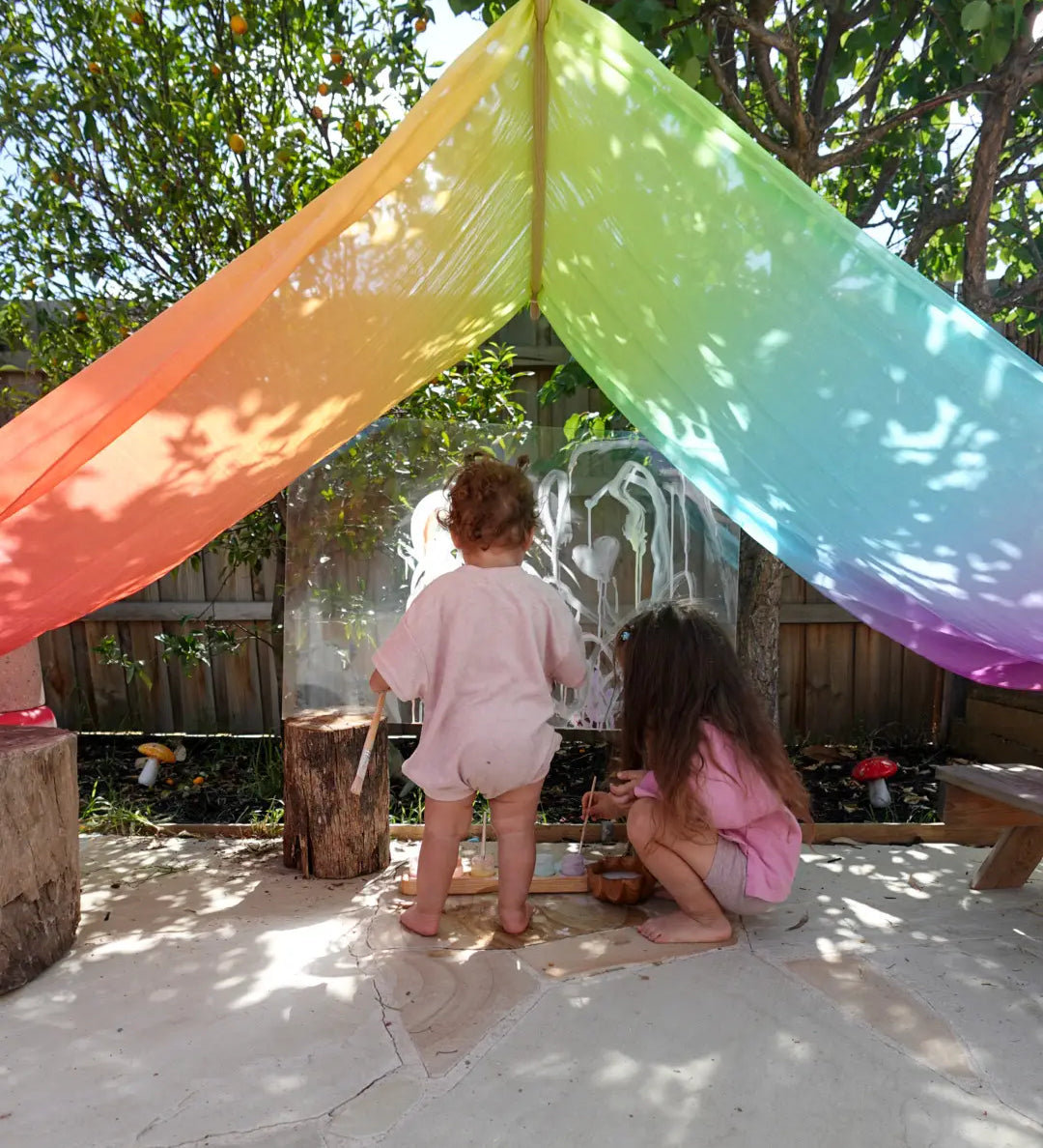 Children playing underneath Laura's play cotton large bright rainbow play cloths in bright rainbow in the garden
