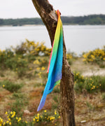 Laura's play cotton bright rainbow play cloths made from organic cotton tied onto a branch with a river in the background