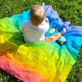 Child sitting on top of Laura's play cotton bright rainbow play cloths on grass