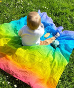 Child sitting on top of Laura's play cotton bright rainbow play cloths on grass