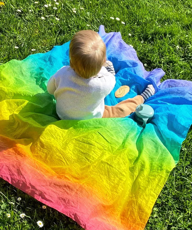 Child sitting on top of Laura's play cotton bright rainbow play cloths on grass