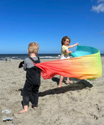 Children playing with Laura's play cotton large pastel rainbow play cloth on the beach