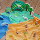 Laura's Play Cotton Play Cloths in blue, green and orange used in a small world play scene with Ostheimer figures and a green Grimm's rainbow in the Babipur playroom.
