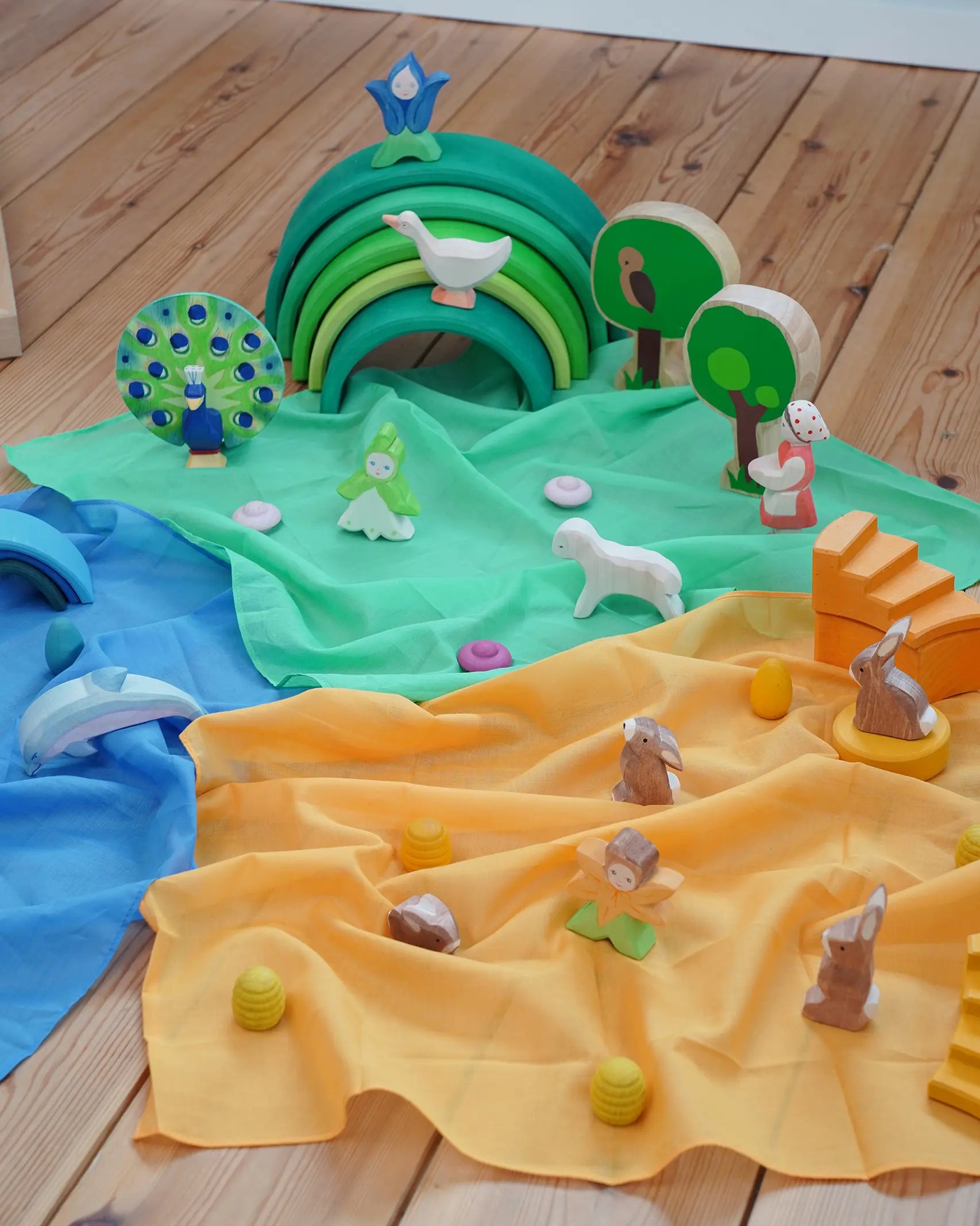 Laura's Play Cotton Play Cloths in blue, green and orange used in a small world play scene with Ostheimer figures and a green Grimm's rainbow in the Babipur playroom.