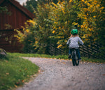 Child riding Early Rider's Belter 16 inch wheel children's bike on a path with trees and a wooden cabin in the background