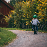 Child riding Early Rider's Belter 16 inch wheel children's bike on a path with trees and a wooden cabin in the background