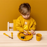 Child wearing Trixie Animal Friends Waterproof Long Sleeve Bib, yellow, Mr Lion, eating from a matching plate and cutlery set. 