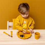 Child wearing Trixie Animal Friends Waterproof Long Sleeve Bib, yellow, Mr Lion, eating from a matching plate and cutlery set. 
